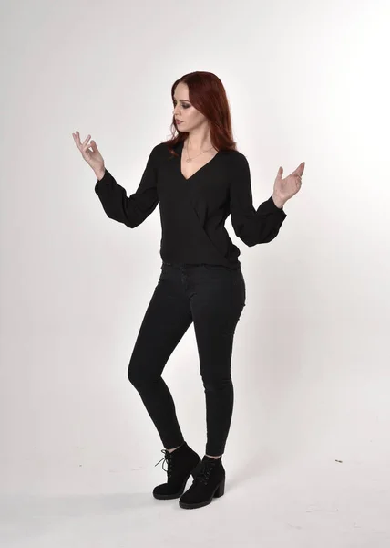 一个红头发 穿着黑色牛仔裤 靴子和衬衫的漂亮女孩的画像 全长站立姿势 在工作室背景上以手势面对镜头 — 图库照片
