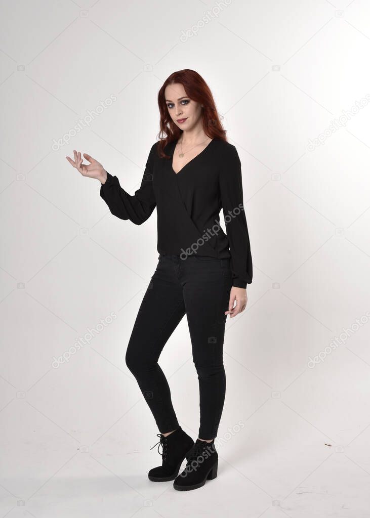 Portrait of a pretty girl with red hair wearing black jeans, boots and a blouse.  full length standing pose, facing the camera with hand gestures on a studio background.