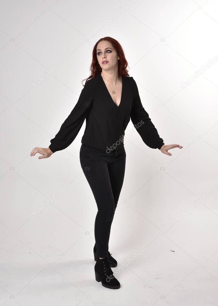 Portrait of a pretty girl with red hair wearing black jeans, boots and a blouse.  full length standing pose, facing the camera with hand gestures on a studio background.