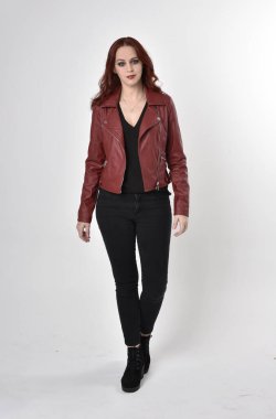 Kızıl saçlı, siyah kot pantolon giyen ve deri ceketli güzel bir kızın portresi. Stüdyo arka planında tam boy ayakta poz.