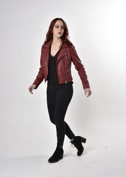 Portrait of a pretty girl with red hair wearing black jeans and boots with leather jacket.  full length standing pose on a studio background.