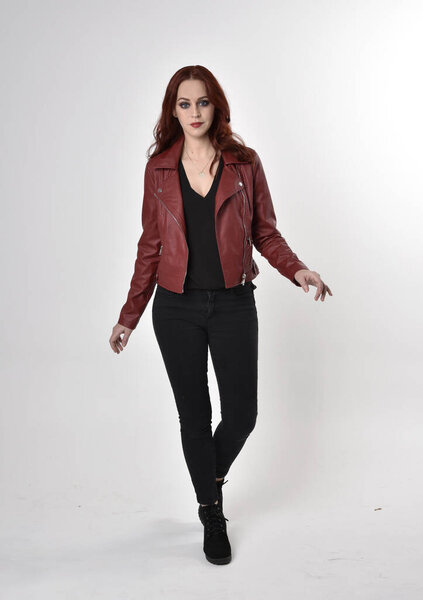 Portrait of a pretty girl with red hair wearing black jeans and boots with leather jacket.  full length standing pose on a studio background.