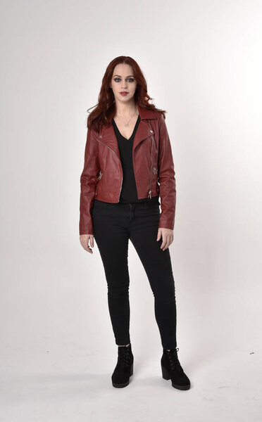 Portrait of a pretty girl with red hair wearing black jeans and boots with leather jacket.  full length standing pose on a studio background.