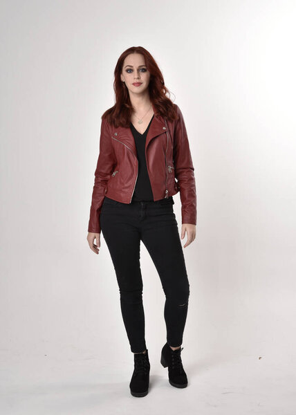 Portrait of a pretty girl with red hair wearing black jeans and boots with leather jacket.  full length standing pose on a studio background.