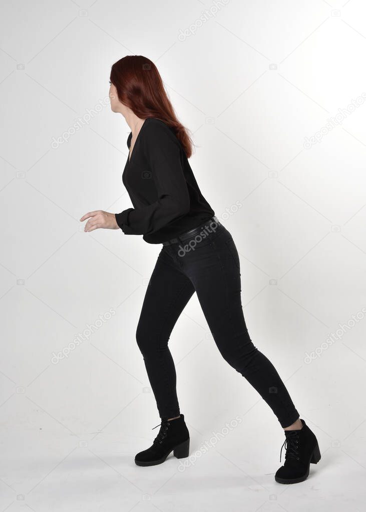 Portrait of a pretty girl with red hair wearing black jeans, boots and a blouse.  full length standing pose in side profile,  on a studio background.