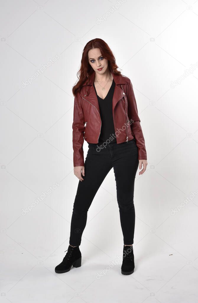 Portrait of a pretty girl with red hair wearing black jeans and boots with leather jacket.  full length standing pose on a studio background.
