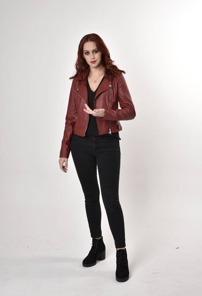 Portrait of a pretty girl with red hair wearing black jeans and boots with leather jacket.  full length standing pose with hand gesture on a studio background.