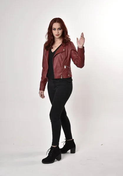 黒のジーンズと革のジャケットとブーツを身に着けている赤い髪のきれいな女の子の肖像画 スタジオの背景に手のジェスチャー付きの全長立ちポーズ — ストック写真