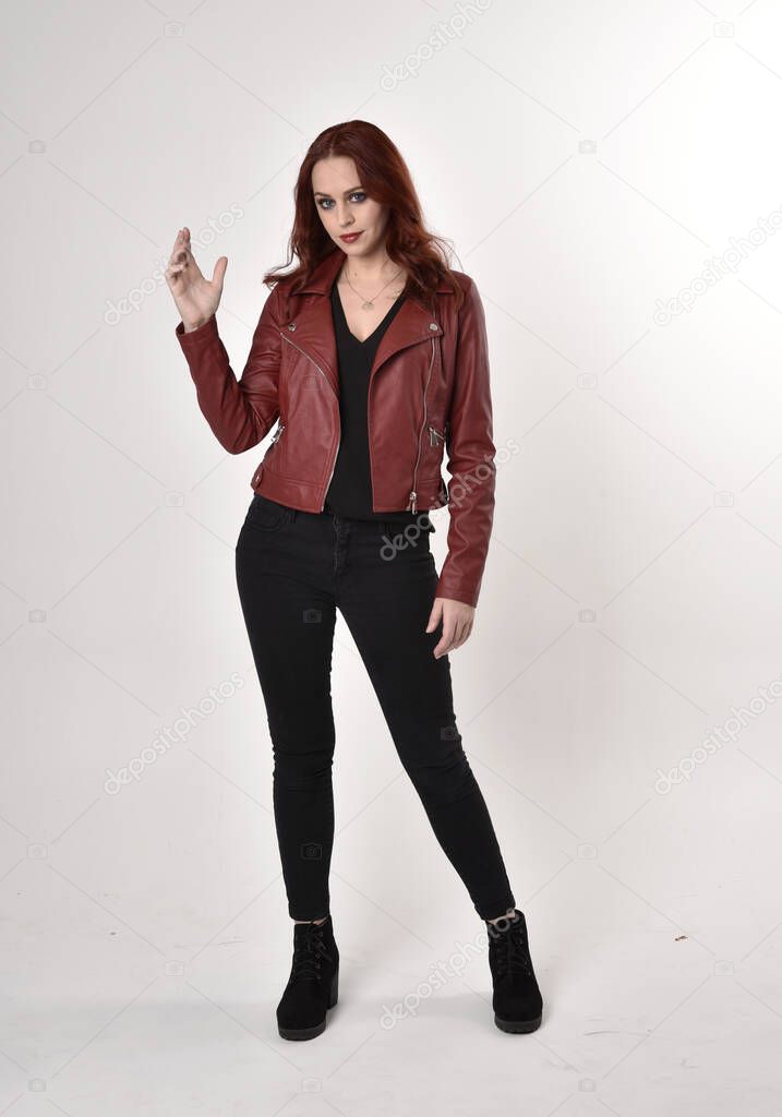 Portrait of a pretty girl with red hair wearing black jeans and boots with leather jacket.  full length standing pose with hand gesture on a studio background.