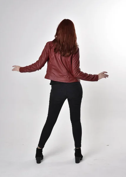 黒のジーンズと革のジャケットとブーツを身に着けている赤い髪のきれいな女の子の肖像画 カメラに背を向けて全長立ちポーズスタジオ背景 — ストック写真