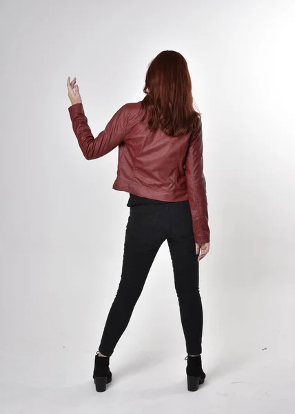 黒のジーンズと革のジャケットとブーツを身に着けている赤い髪のきれいな女の子の肖像画 カメラに背を向けて全長立ちポーズスタジオ背景 — ストック写真