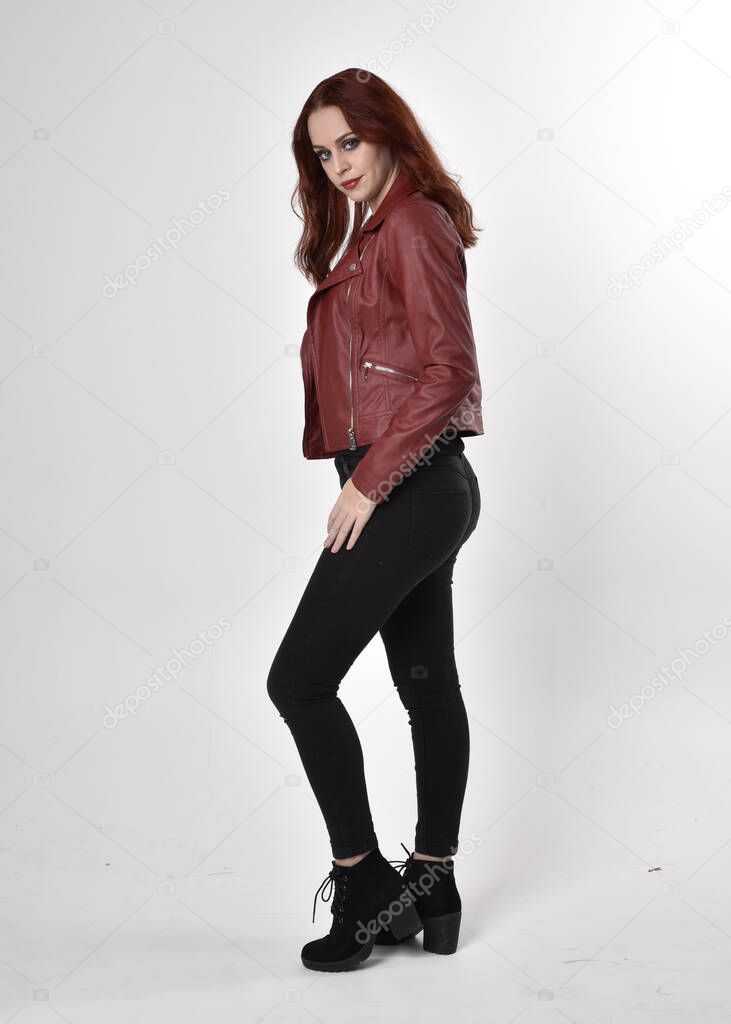 Portrait of a pretty girl with red hair wearing black jeans and boots with leather jacket.  full length standing pose with back to the camera a studio background.