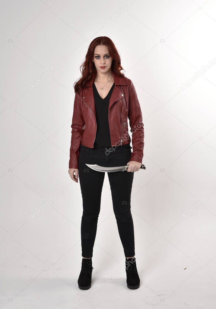 Portrait of a pretty girl with red hair wearing black jeans and boots with leather jacket.  full length standing pose with back to the camera a studio background.