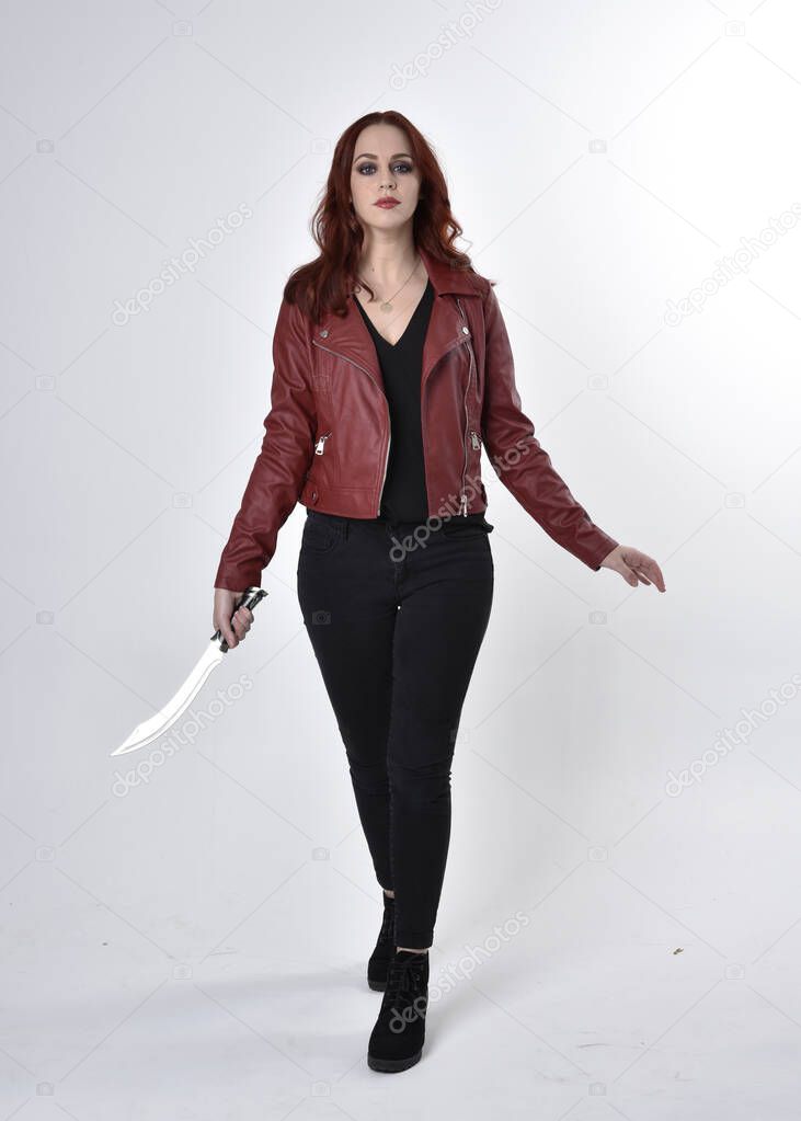 Portrait of a pretty girl with red hair wearing black jeans and boots with leather jacket.  full length standing pose holding knife a studio background.