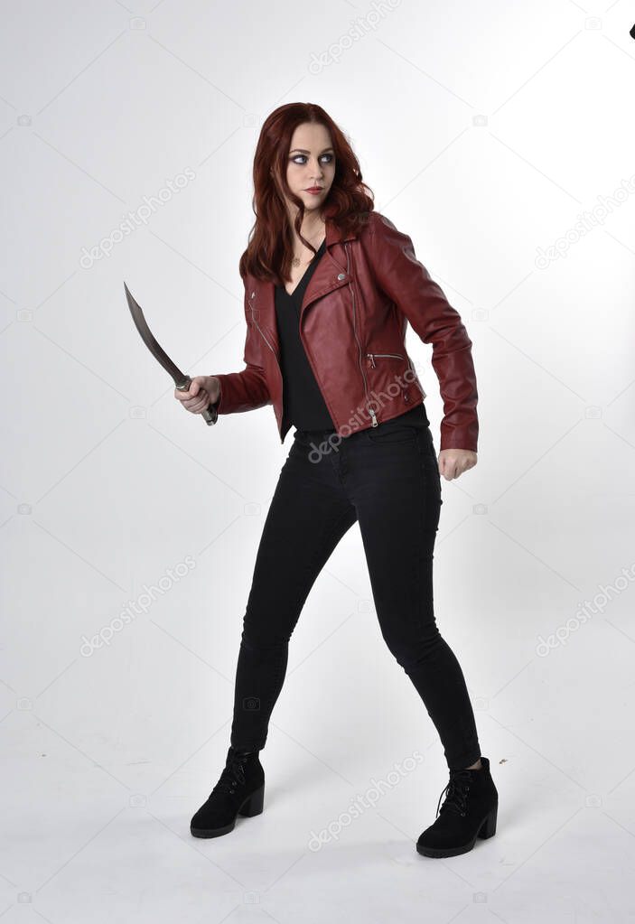 Portrait of a pretty girl with red hair wearing black jeans and boots with leather jacket.  full length standing pose holding knife a studio background.