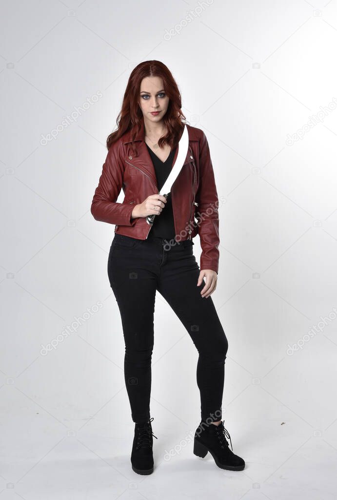 Portrait of a pretty girl with red hair wearing black jeans and boots with leather jacket.  full length standing pose holding knife a studio background.
