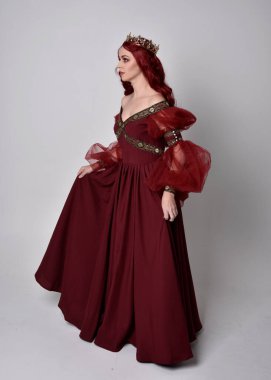 Havada uçuşan Burgundy fantezi elbisesi ve altın taç giyen kızıl saçlı güzel bir kadının portresi. Tam boy ayakta duruş, stüdyo arka planında izole edilmiş.