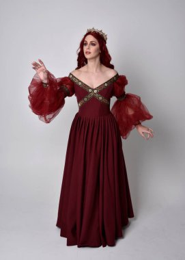 Havada uçuşan Burgundy fantezi elbisesi ve altın taç giyen kızıl saçlı güzel bir kadının portresi. Tam boy ayakta duruş, stüdyo arka planında izole edilmiş.