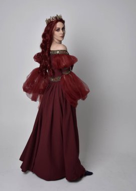 Havada uçuşan Burgundy fantezi elbisesi ve altın taç giyen kızıl saçlı güzel bir kadının portresi. Tam boy ayakta duruş, stüdyo arka planında izole edilmiş.