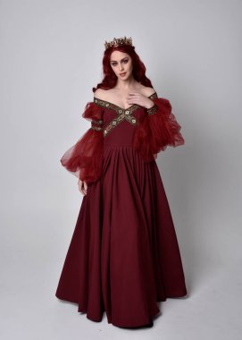 Havada uçuşan Burgundy fantezi elbisesi ve altın taç giyen kızıl saçlı güzel bir kadının portresi. Tam boy ayakta duruş, stüdyo arka planında izole edilmiş.