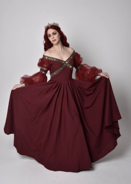 Havada uçuşan Burgundy fantezi elbisesi ve altın taç giyen kızıl saçlı güzel bir kadının portresi. Tam boy ayakta duruş, stüdyo arka planında izole edilmiş.