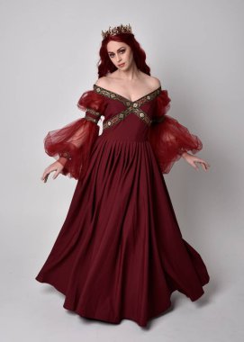 Havada uçuşan Burgundy fantezi elbisesi ve altın taç giyen kızıl saçlı güzel bir kadının portresi. Tam boy ayakta duruş, stüdyo arka planında izole edilmiş.