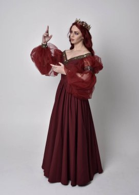 Havada uçuşan Burgundy fantezi elbisesi ve altın taç giyen kızıl saçlı güzel bir kadının portresi. Tam boy ayakta duruş, stüdyo arka planında izole edilmiş.