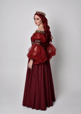 Havada uçuşan Burgundy fantezi elbisesi ve altın taç giyen kızıl saçlı güzel bir kadının portresi. Tam boy ayakta duruş, stüdyo arka planında izole edilmiş.