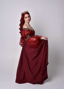 Havada uçuşan Burgundy fantezi elbisesi ve altın taç giyen kızıl saçlı güzel bir kadının portresi. Tam boy ayakta duruş, stüdyo arka planında izole edilmiş.