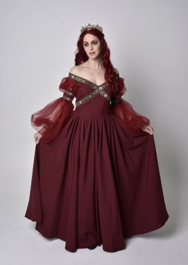 Havada uçuşan Burgundy fantezi elbisesi ve altın taç giyen kızıl saçlı güzel bir kadının portresi. Tam boy ayakta duruş, stüdyo arka planında izole edilmiş.