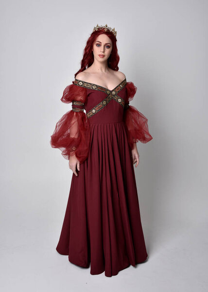 Портрет красивой женщины с рыжими волосами в бургундском фэнтези-платье и золотой короне. полностью стоящая поза, изолированная от студийного фона
