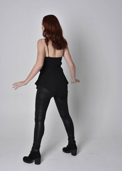  portrait of a pretty girl with red hair wearing black leather pants and top. Full length standing pose with back to the camera  isolated against a  grey studio background