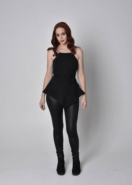 portrait of a pretty girl with red hair wearing black leather pants and top. Full length standing pose isolated against a  grey studio background