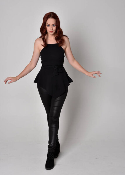 portrait of a pretty girl with red hair wearing black leather pants and top. Full length standing pose isolated against a  grey studio background