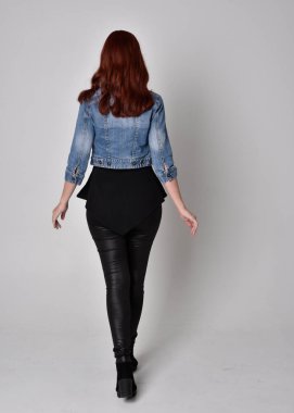 Siyah deri pantolon ve kot ceket giyen kızıl saçlı güzel bir kızın portresi. Gri bir stüdyo arka planında tam boy ayakta poz