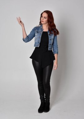  Siyah deri pantolon ve kot ceket giyen kızıl saçlı güzel bir kızın portresi. Gri bir stüdyo arka planında tam boy ayakta poz