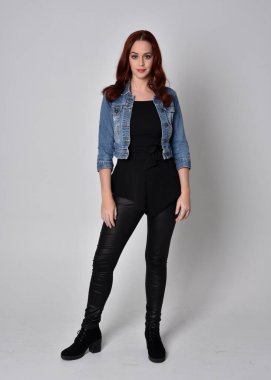  Siyah deri pantolon ve kot ceket giyen kızıl saçlı güzel bir kızın portresi. Gri bir stüdyo arka planında tam boy ayakta poz