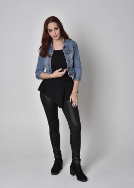  Siyah deri pantolon ve kot ceket giyen kızıl saçlı güzel bir kızın portresi. Gri bir stüdyo arka planında tam boy ayakta poz