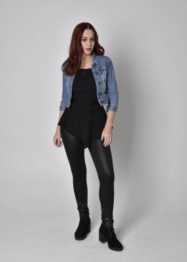  Siyah deri pantolon ve kot ceket giyen kızıl saçlı güzel bir kızın portresi. Gri bir stüdyo arka planında tam boy ayakta poz