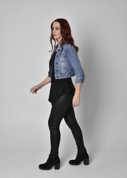 portrait of a pretty girl with red hair wearing black leather pants and denim jacket. Full length standing pose isolated against a  grey studio background