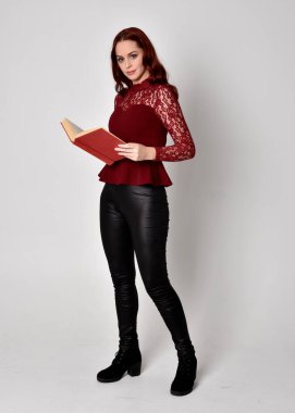 Deri pantolon ve uzun kollu gömlek giyen kızıl saçlı güzel bir kızın portresi. Stüdyonun arka planında izole bir şekilde duran bir kitap tutuyor..