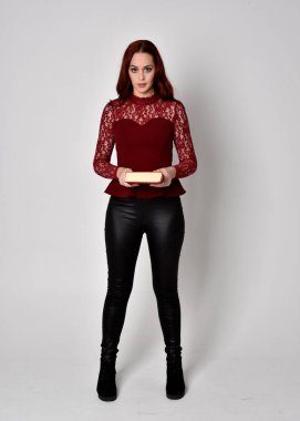 Deri pantolon ve uzun kollu gömlek giyen kızıl saçlı güzel bir kızın portresi. Stüdyonun arka planında izole bir şekilde duran bir kitap tutuyor..