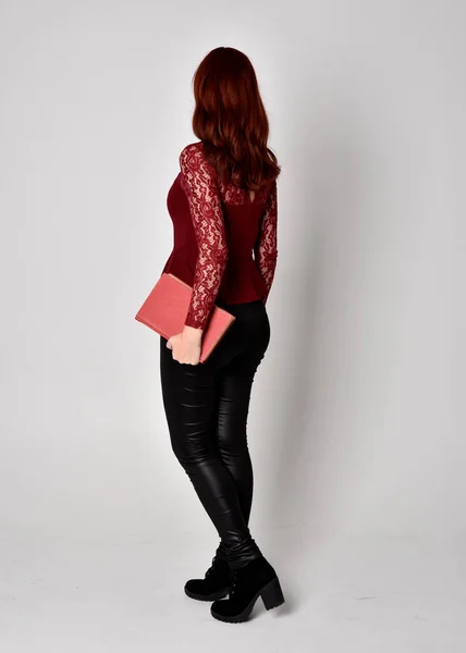 一个穿着皮裤和长袖花边衬衫的红头发美女的画像 全身而立 拿着一本书 与工作室背景隔离 — 图库照片