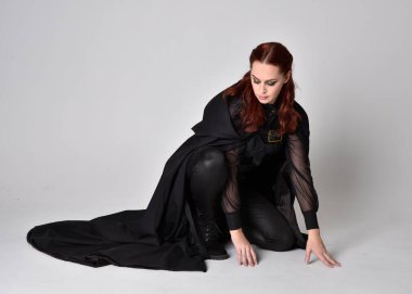 Kızıl saçlı bir kadının fantezi portresi. Uzun siyah pelerinli koyu deri bir suikastçı kostümü giyiyor. Tam boy diz çökme pozisyonu, stüdyo arka planında izole edilmiş..