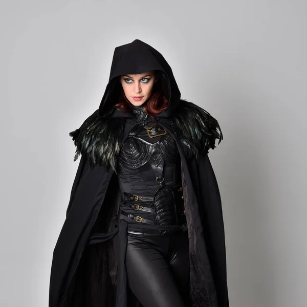 Close up fantasy portrait of a woman with red hair wearing dark leather assassin costume with long black cloak.  isolated against a studio background.