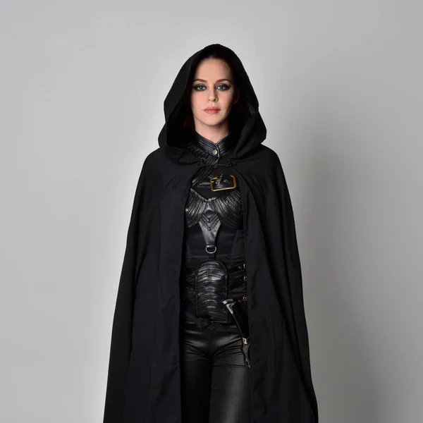 Close up fantasy portrait of a woman with red hair wearing dark leather assassin costume with long black cloak.  isolated against a studio background.