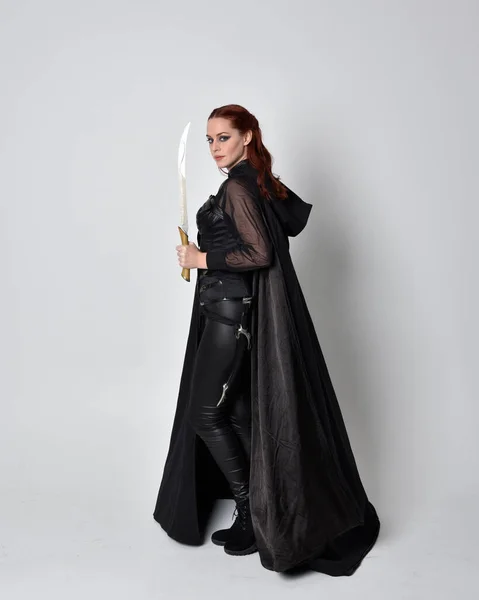 fantasy portrait of a woman with red hair wearing dark leather assassin costume with long black cloak. Full length standing pose holding a sword isolated against a studio background.