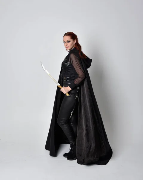 一个红头发的女人穿着黑皮刺客服装和黑色长斗篷的幻想画像 全身而立 拿着剑与工作室背景隔离 — 图库照片