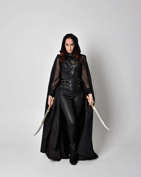 一个红头发的女人穿着黑皮刺客服装和黑色长斗篷的幻想画像 全身而立 拿着剑与工作室背景隔离 — 图库照片