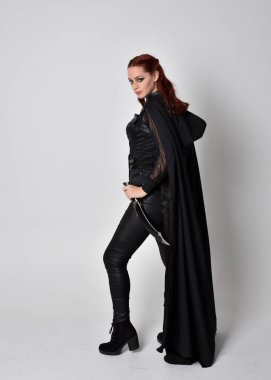 Kızıl saçlı bir kadının fantezi portresi. Uzun siyah pelerinli koyu deri bir suikastçı kostümü giyiyor. Stüdyonun arka planında izole edilmiş tam boy yan profil pozu.